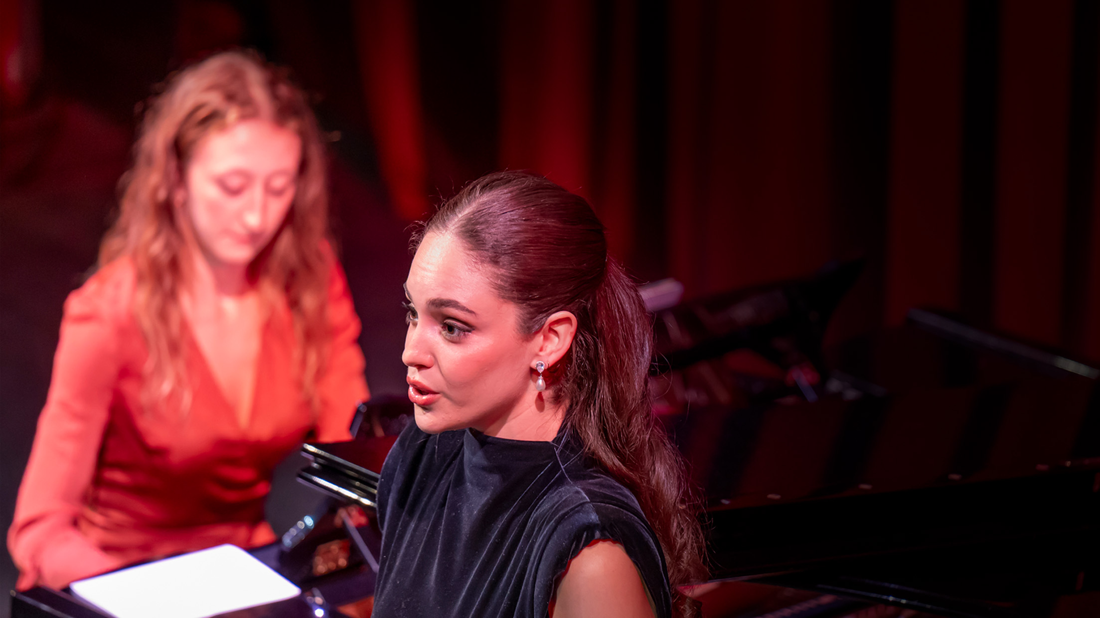 "Your time to shine". A singer wearing a black dress, performing on stage, with a pianist accompanying them on stage in an orange dress.